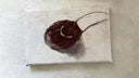 A flat lay photograph of a finished handbag painted on a small canvas, placed on a textured beige surface. The canvas features a dark brown painted handbag with a white curved stripe.
