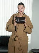 Reco. Casa rosewood leather baguette bag with adjustable strap and key charm detail. Model wears camel coat.
