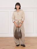 A model wearing a beige zip-up sweater and wide-leg trousers holding a brown Reco. Halston crescent-shaped Togo calfskin bag.