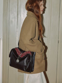 Reco. Rosewood leather shoulder bag with compass charm and red knit detail, worn by a woman in a tan coat.