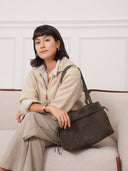 Model wearing a Reco. L'erreur dumpling bag in Falcon Gray nubuck leather with a drawstring top on a beige sofa.